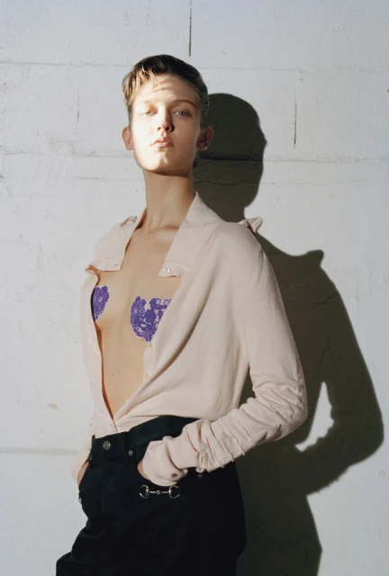 A young adult woman poses indoors wearing a light pink collared shirt, black pants, and purple heart-shaped pasties against a brick wall.