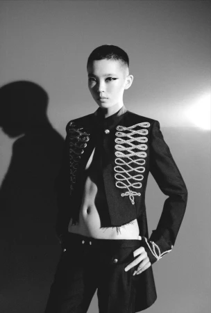 Monochrome portrait of a person with a shaved head, wearing a military-style jacket with elaborate frogging and black trousers in studio lighting.