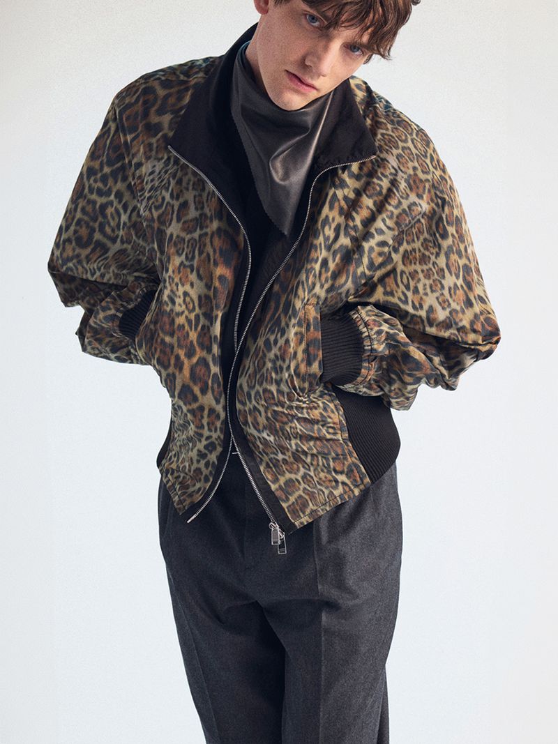 Portrait of a young adult male wearing a leopard print jacket, grey pants, and dark shirt, posing indoors against a plain light-colored background.
