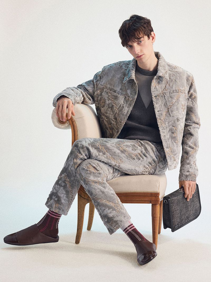 Male model seated in an upholstered chair wearing a grey patterned jacket and trousers, holding a dark clutch against a white seamless backdrop.