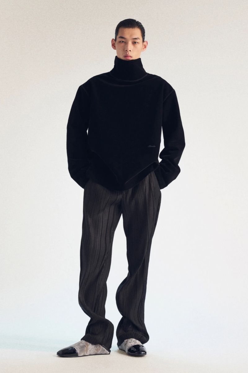 Full body studio shot of an East Asian male model wearing a black turtleneck sweater, black pleated pants, and black and silver shoes.