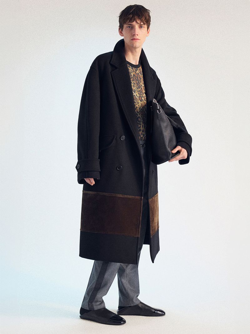 A fair-skinned adult male model poses in a black double-breasted coat with brown detailing, gray trousers, and black flats in a studio setting.