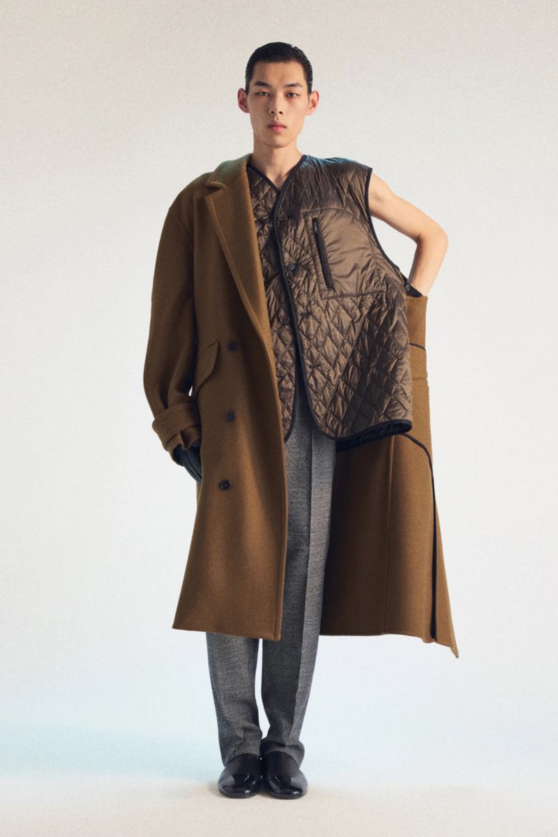 Editorial fashion shot of a slender Asian man wearing a brown overcoat, quilted vest, patterned shirt, and grey trousers against a white backdrop.
