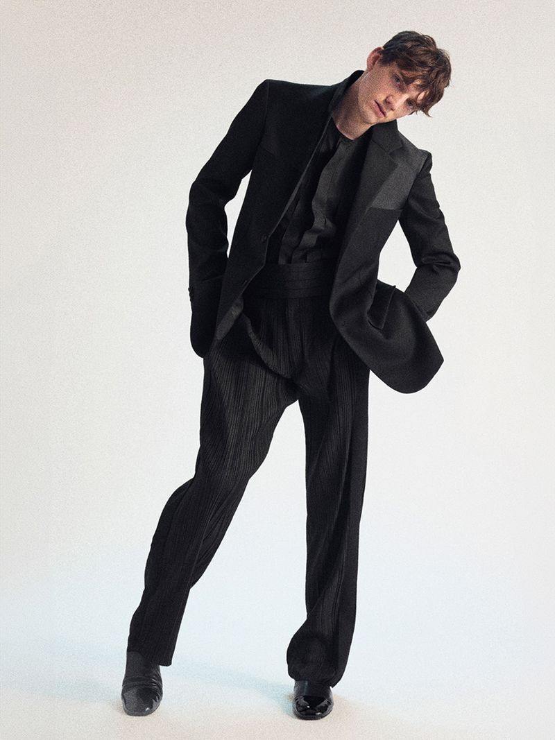 Male fashion model in all-black suit strikes a pose in a full shot against a clean white backdrop. He wears a dark shirt, pleated pants, and shiny shoes.