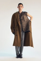 Model wearing a brown coat draped over one shoulder and a quilted vest over grey trousers against a plain white background