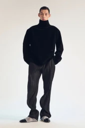 Model wearing a black turtleneck sweater and pleated trousers against a neutral white background.