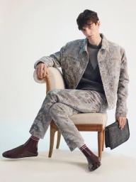 Male model seated in an upholstered chair wearing a grey patterned jacket and trousers, holding a dark clutch against a white seamless backdrop.