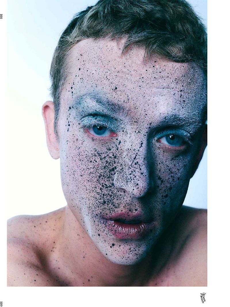Close-up portrait of a young man with light skin, blue eyes, and short brown hair, wearing pale makeup with black speckled details.
