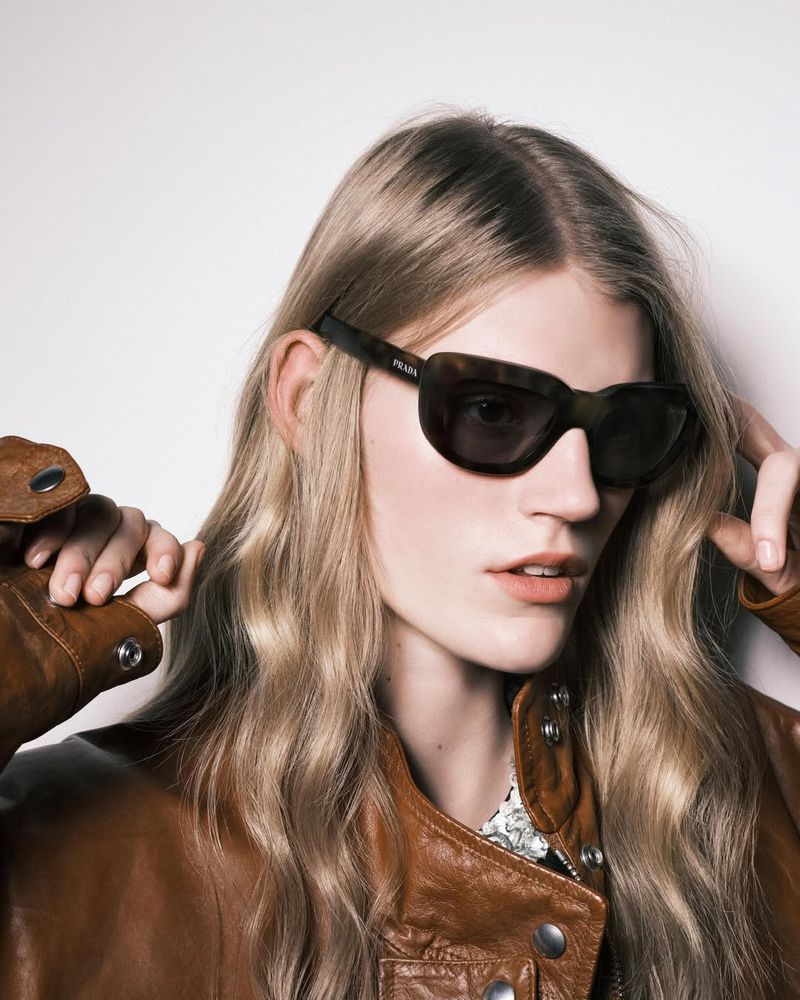 A woman with long blonde hair wears tortoiseshell Prada sunglasses and a brown leather jacket against a plain light background.