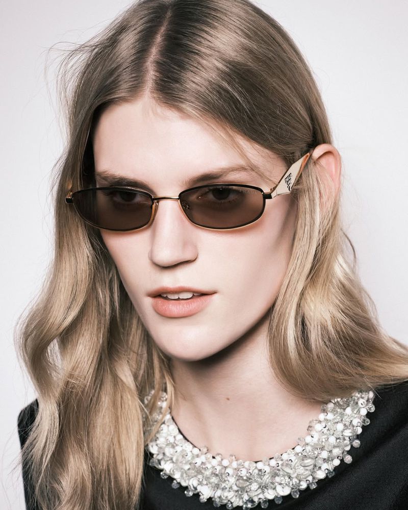 A blonde woman with sunglasses and a beaded collar poses in a close-up shot. The image focuses on fashion and beauty.