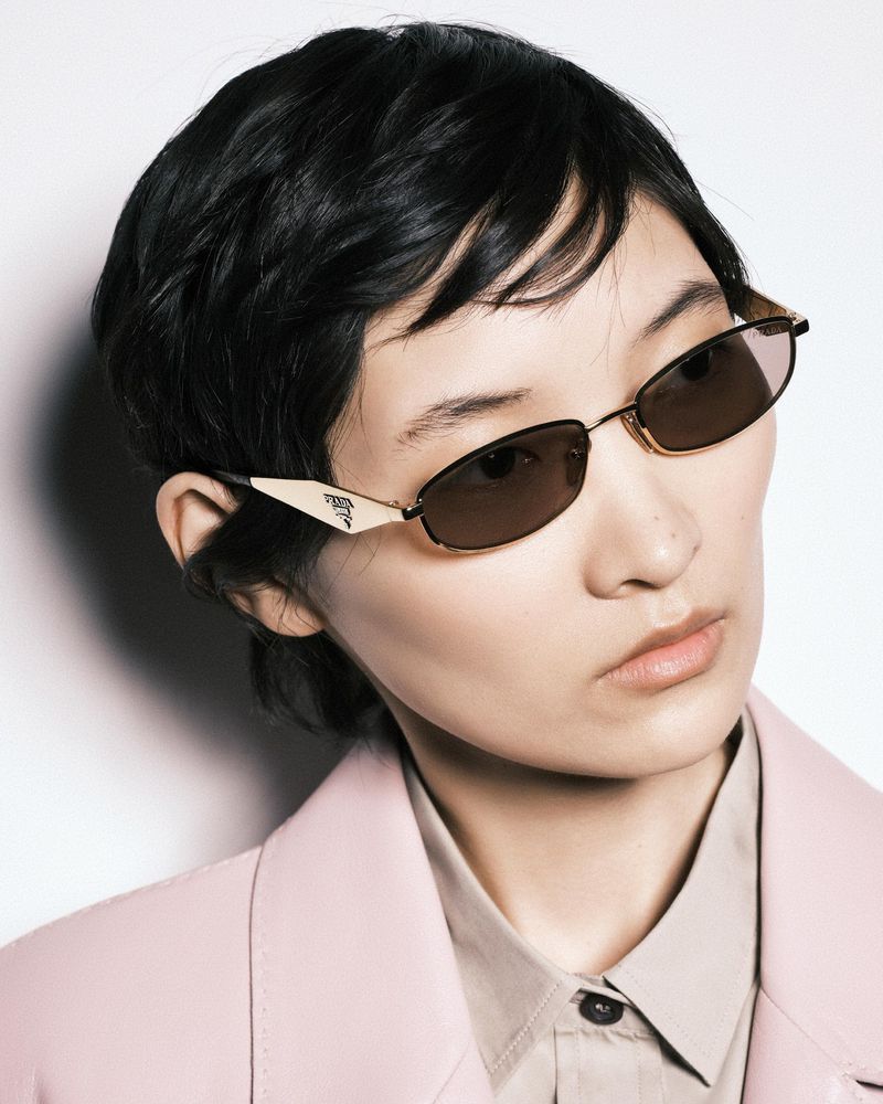 Close-up of an Asian woman wearing a light pink jacket, beige button-up shirt, and stylish Prada sunglasses against a white background.