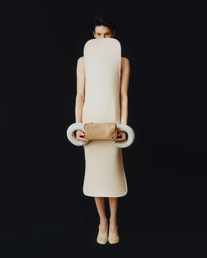 Full shot of a woman in an ivory dress and heels, holding a small bag with sculptural rings, against a black studio backdrop.