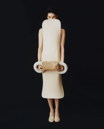 Full shot of a woman in an ivory dress and heels, holding a small bag with sculptural rings, against a black studio backdrop.