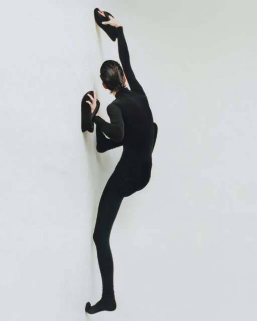 Athletic person in a black bodysuit is posing on a white wall, legs and arms extended in an artistic gymnastic form.