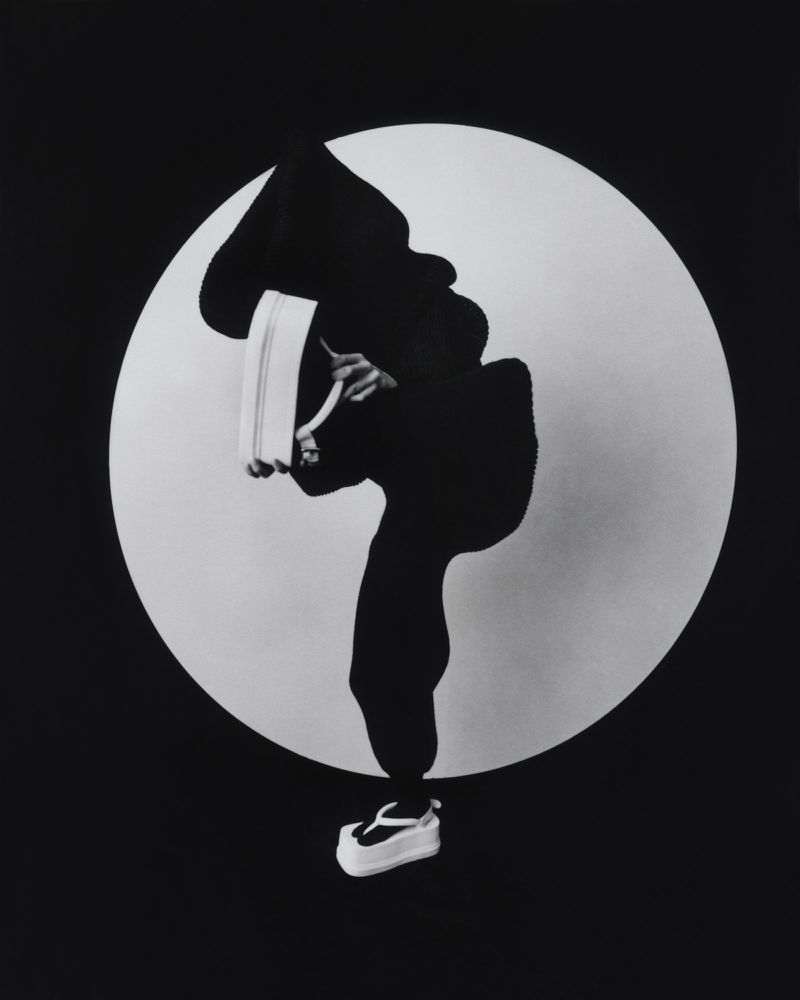 Person in black outfit strikes dynamic pose against white circle and black background, holding a white object and wearing platform sandals.