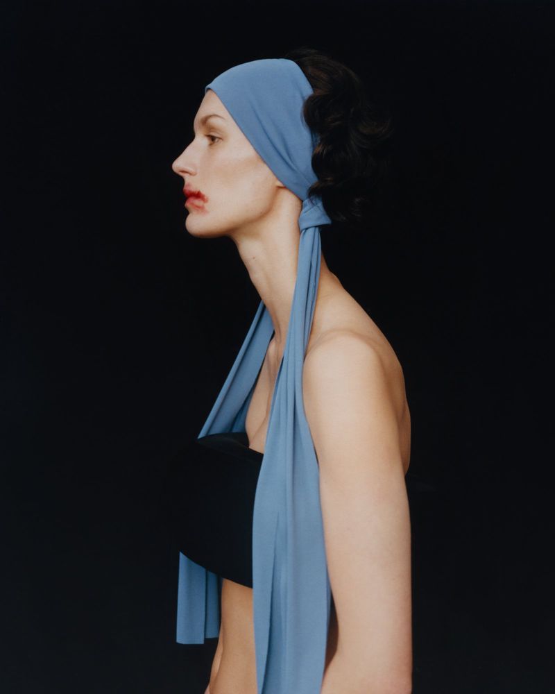Profile shot of a woman with dark hair wearing a light blue headscarf and black top against a dark background in a studio.