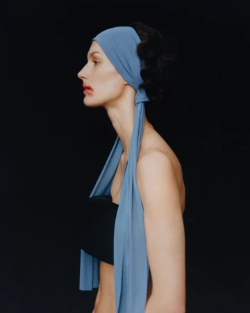 Profile shot of a woman with dark hair wearing a light blue headscarf and black top against a dark background in a studio.
