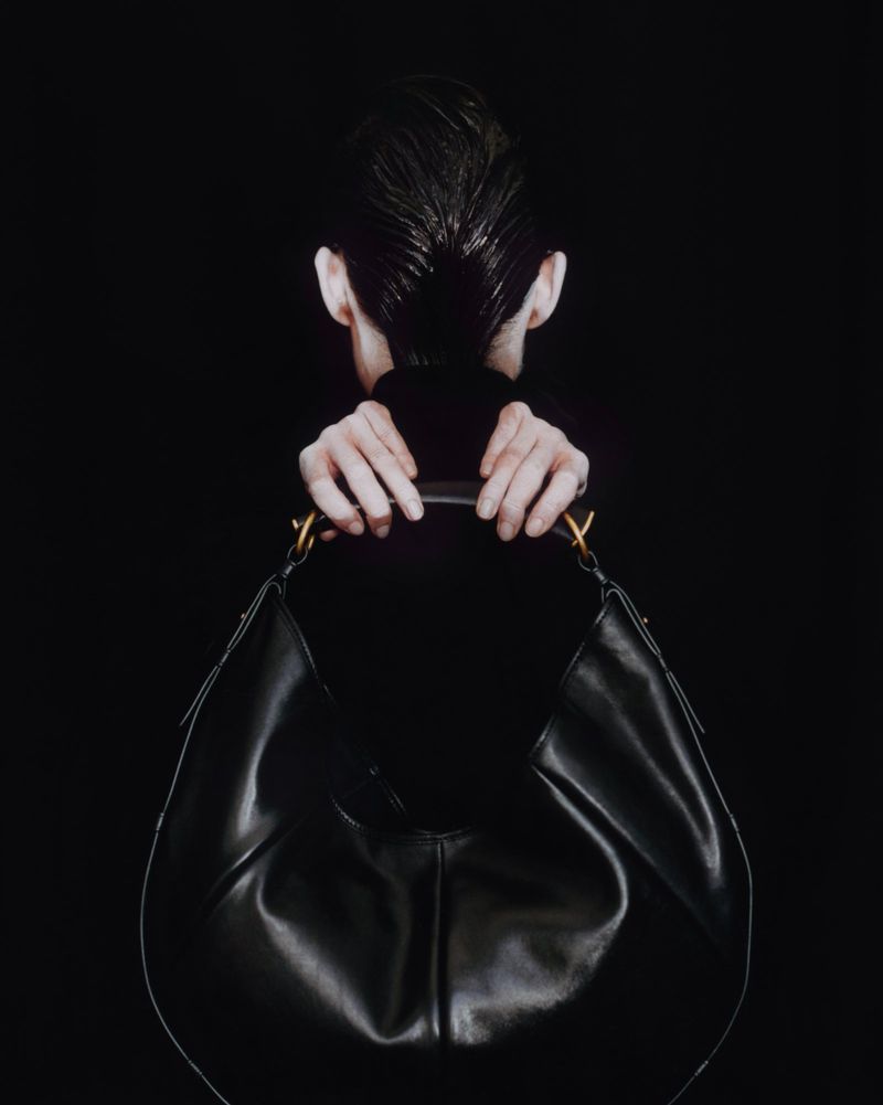 Fashion shot of an unrecognizable model holding a large black leather hobo bag with gold handle detail against a dark background.