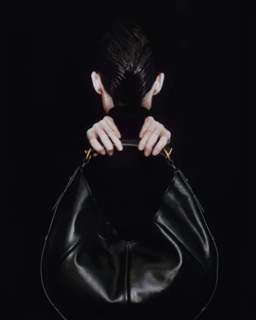 Fashion shot of an unrecognizable model holding a large black leather hobo bag with gold handle detail against a dark background.