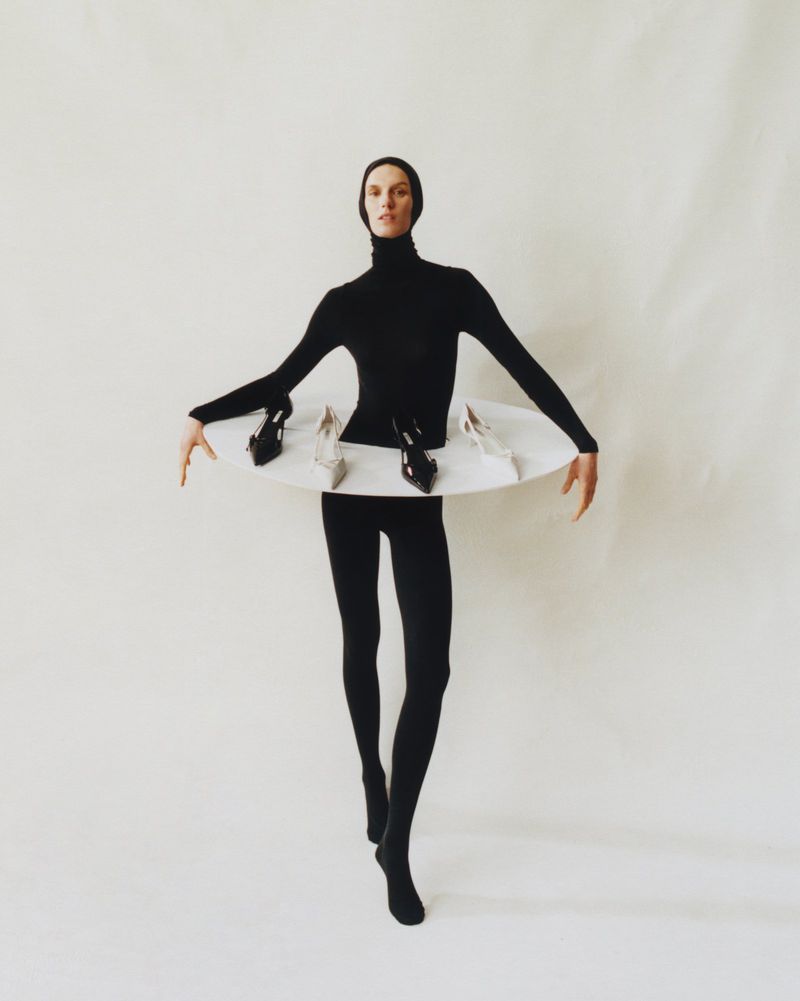Woman in black bodysuit strikes a pose with a white disk around her waist holding several high heel shoes against a white backdrop.