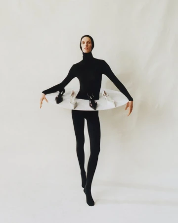 Woman in black bodysuit strikes a pose with a white disk around her waist holding several high heel shoes against a white backdrop.