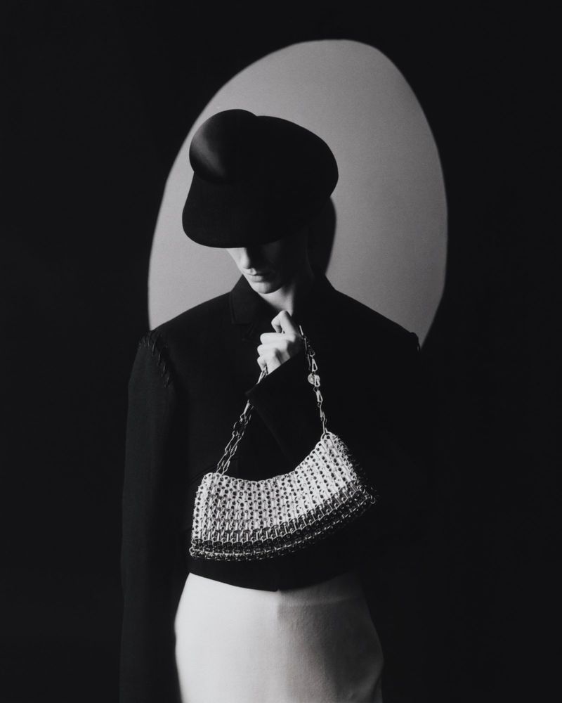 Monochrome portrait of a woman in a black hat and jacket, holding a silver handbag against a dark background with a soft light.