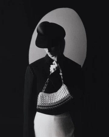 Monochrome portrait of a woman in a black hat and jacket, holding a silver handbag against a dark background with a soft light.