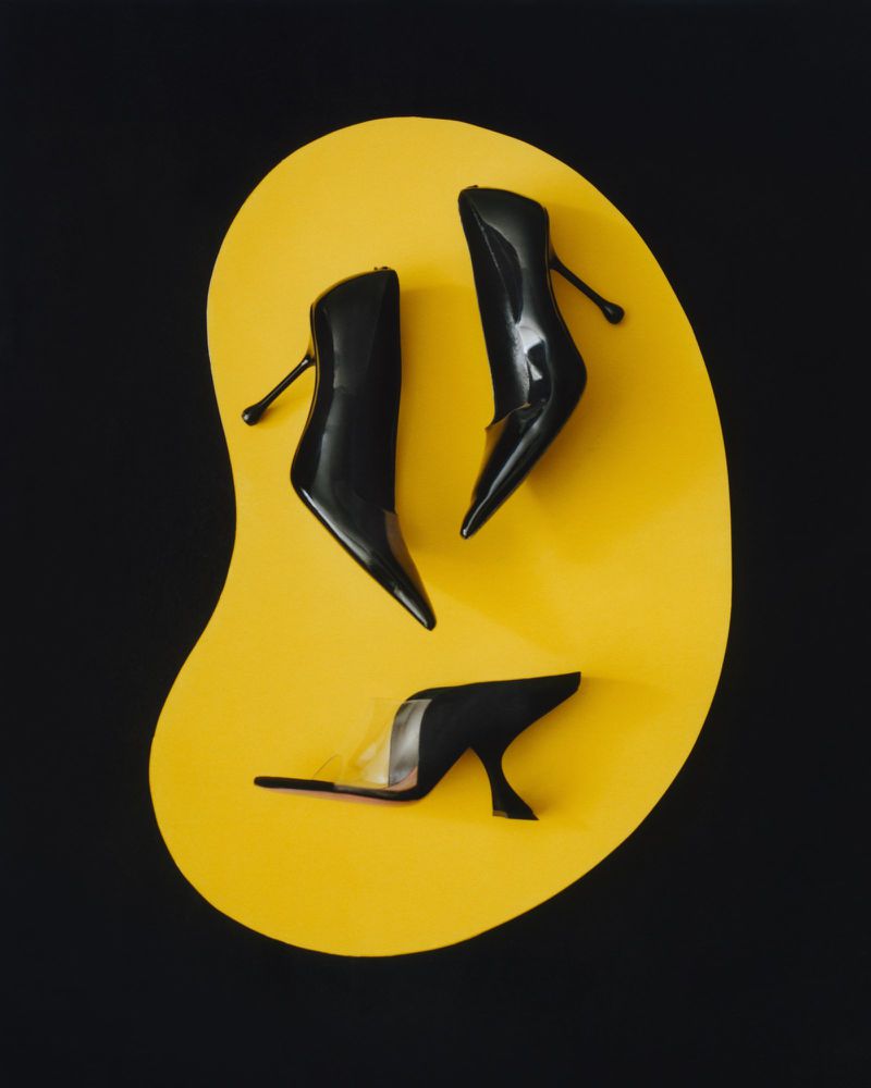 Three black stiletto heels are artfully arranged on a smooth, kidney-shaped yellow platform against a stark black backdrop, in studio lighting.