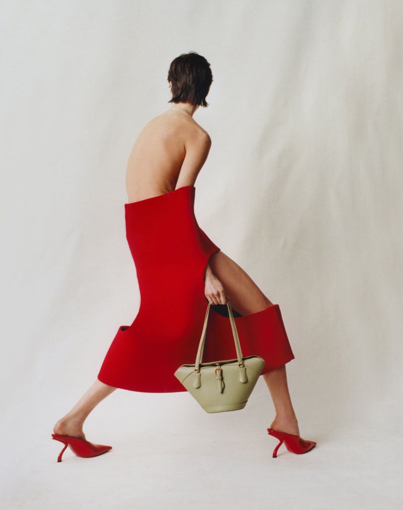 A bare-backed woman in a red dress and red heels strides across a light background while holding a light green handbag.