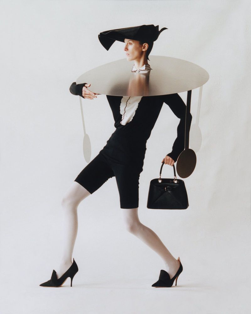 A person models avant-garde fashion, including a wide-brimmed hat, metallic collar, and black handbag, against a bright white backdrop in a studio.