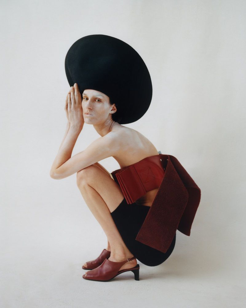Woman with painted face crouches wearing large black hat and maroon fashion corset in a studio setting with white backdrop.