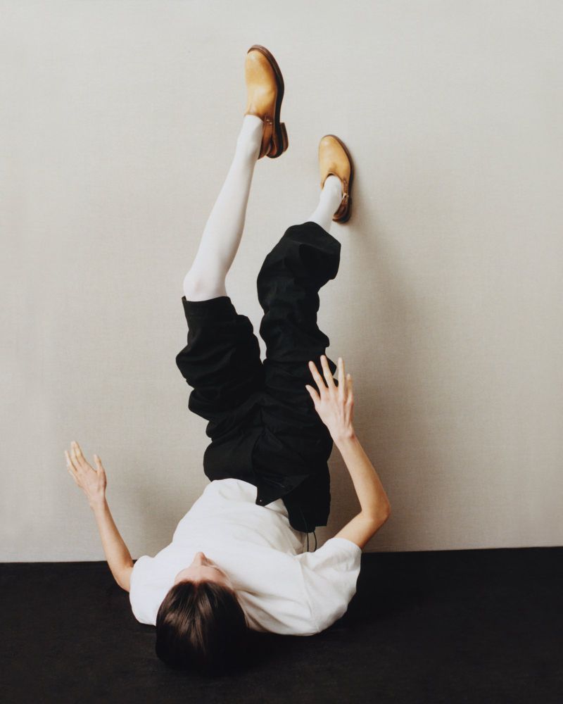 Upside down view of a person against a wall wearing a white t-shirt, black pants, white socks and brown shoes.
