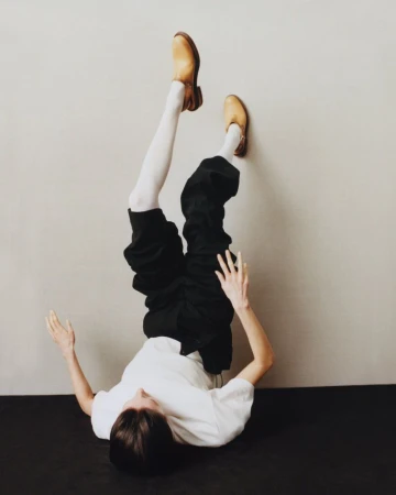 Upside down view of a person against a wall wearing a white t-shirt, black pants, white socks and brown shoes.