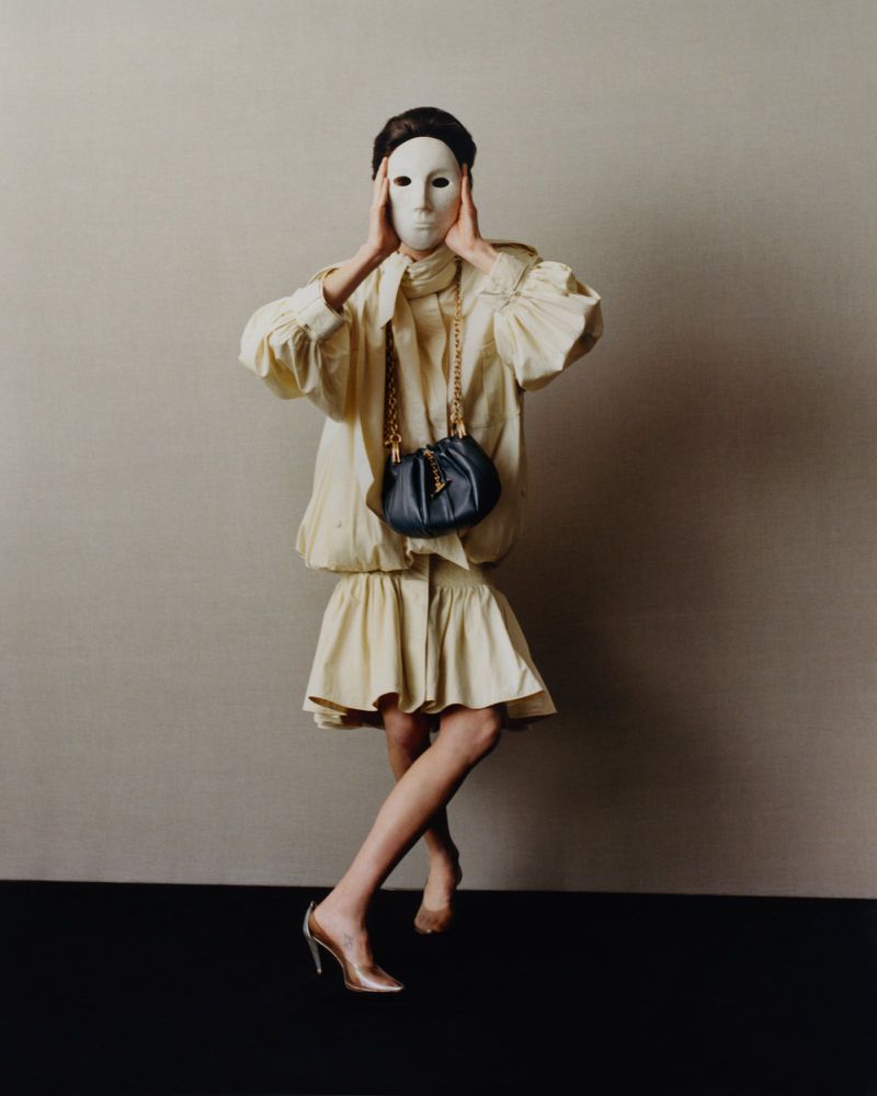 A person wearing a cream-colored dress, covered by a white mask, poses in front of a neutral wall with a dark purse and heels.