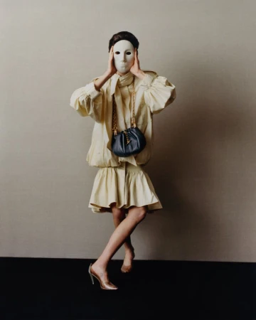 A person wearing a cream-colored dress, covered by a white mask, poses in front of a neutral wall with a dark purse and heels.