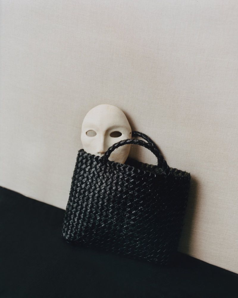 A black woven tote bag sits on a dark surface with a white mask partially inside against a beige wall, creating a minimalist composition.