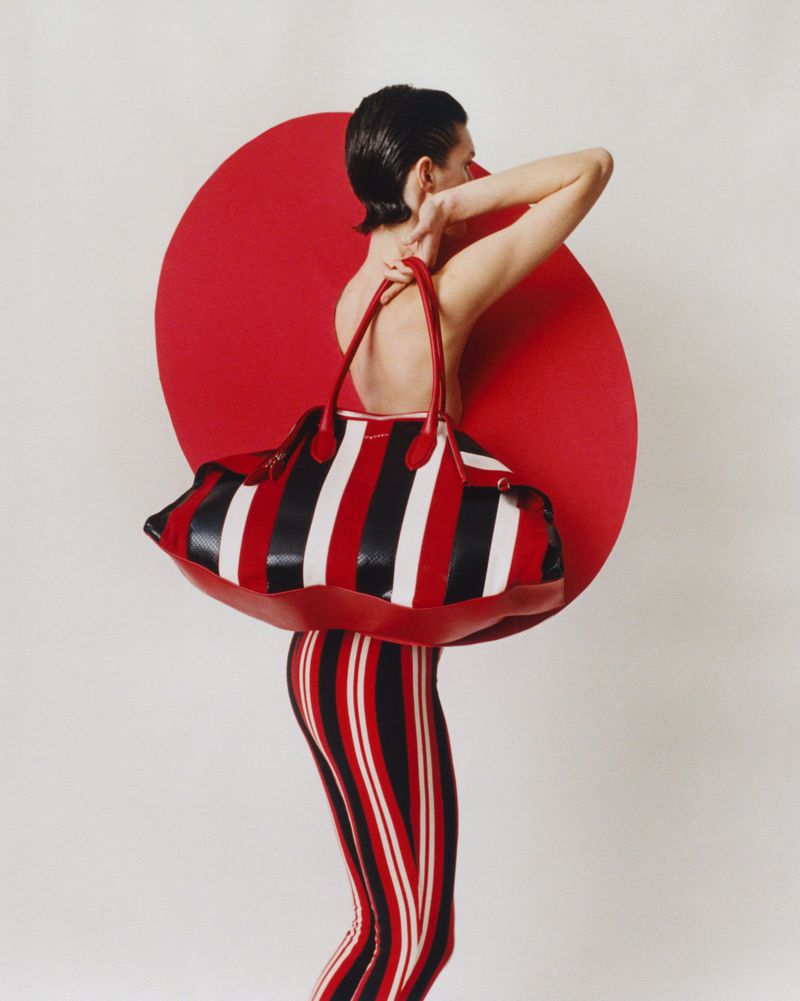 Back view of a model with short brown hair holding a striped duffel bag in front of a red circle on a white background.