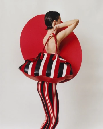 Back view of a model with short brown hair holding a striped duffel bag in front of a red circle on a white background.