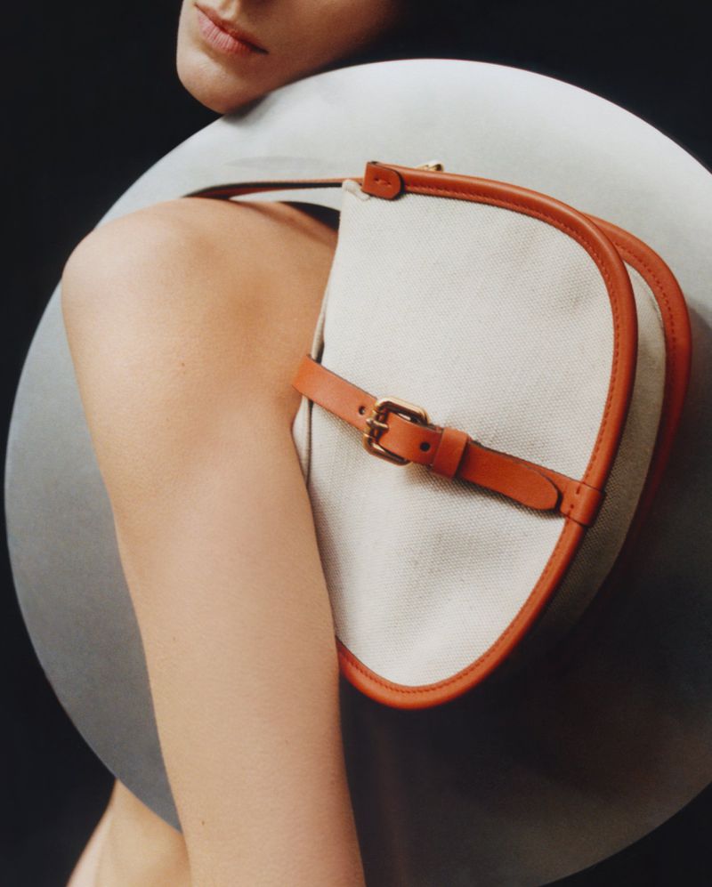 Close-up of a light-skinned woman's shoulder and part of her face, with a beige handbag with tan leather straps against a dark background.