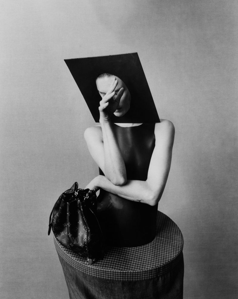 Black and white fashion shot of a person with hand to face, wearing a geometric hat and dress, seated next to a patterned purse.