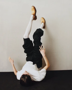 Model lying upside down against a neutral wall wearing a white t-shirt, black pants, white tights, and tan shoes.