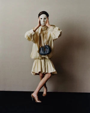 Model wearing a white mask, light-colored puffy dress, and dark shoulder bag holding hands to the mask against a neutral studio backdrop