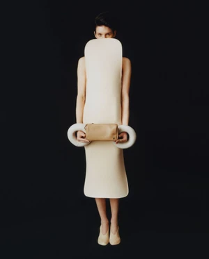 Model Marte Mei van Haaster wearing a sculptural cream garment holding a beige clutch against a black background