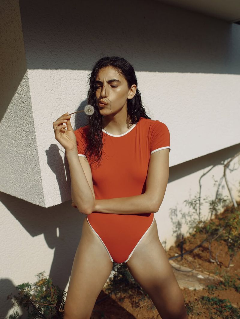 Adult female in a red bodysuit posing with a dandelion in an outdoor setting with greenery and a white wall backdrop.