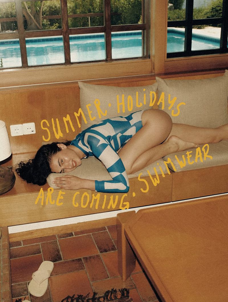 Brunette woman in patterned swimsuit relaxes on sofa by a window overlooking a pool, evoking a summery and relaxed lifestyle mood.