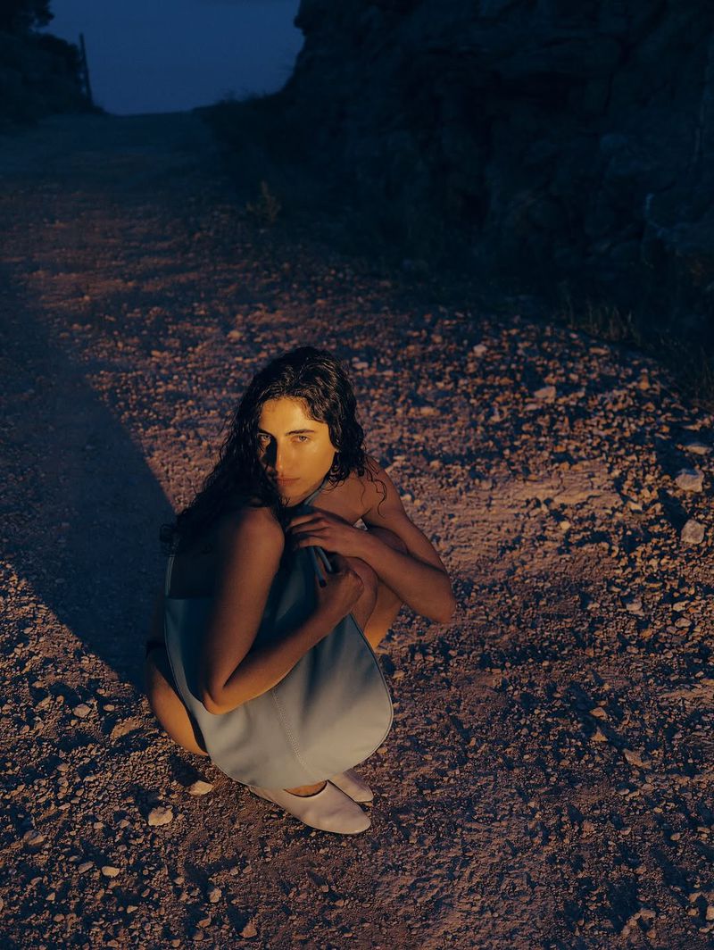 Brunette woman in blue dress squats on rocky ground in low light. She looks directly at the camera in an outdoor setting.
