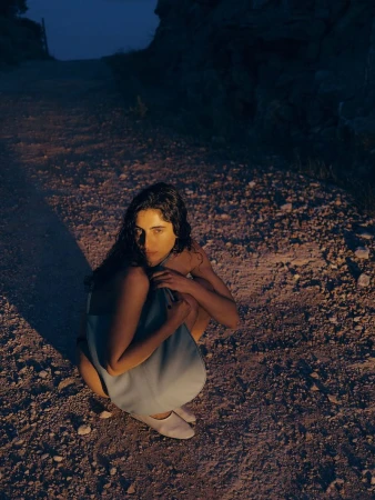 Brunette woman in blue dress squats on rocky ground in low light. She looks directly at the camera in an outdoor setting.