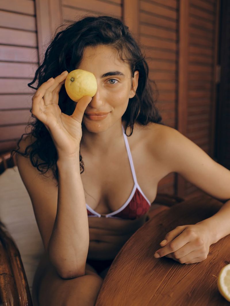 Portrait of a woman holding a lemon up to her eye while sitting at a table in a red bikini top with a neutral expression.