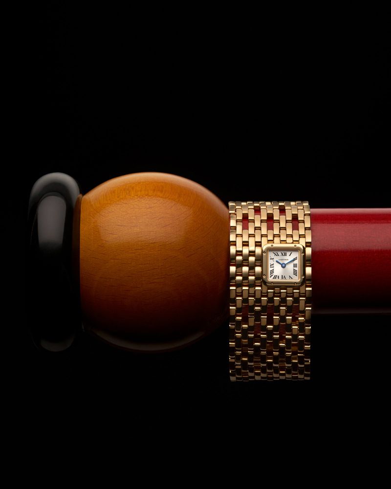 Close-up of a gold wristwatch with a square face wrapped around a brown and red object, set against a stark black background.