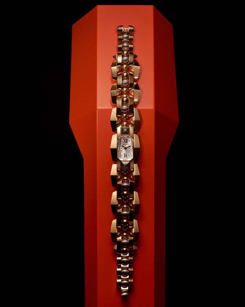 Elevated view of a gold bracelet watch with a geometric band, presented on a red display pedestal against a dark background.